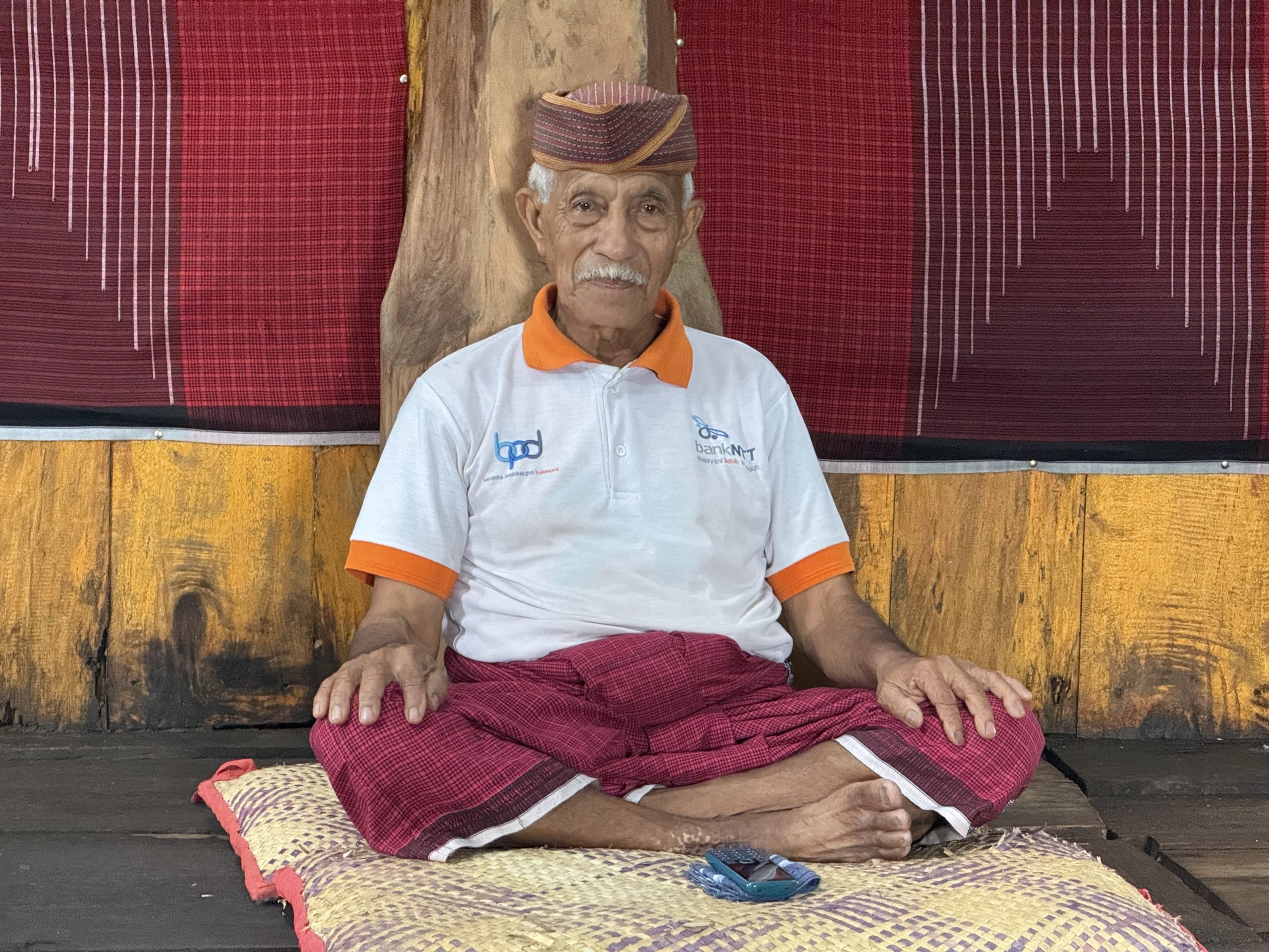 An elder sitting cross-legged on a mat inside a traditional Manggarai house