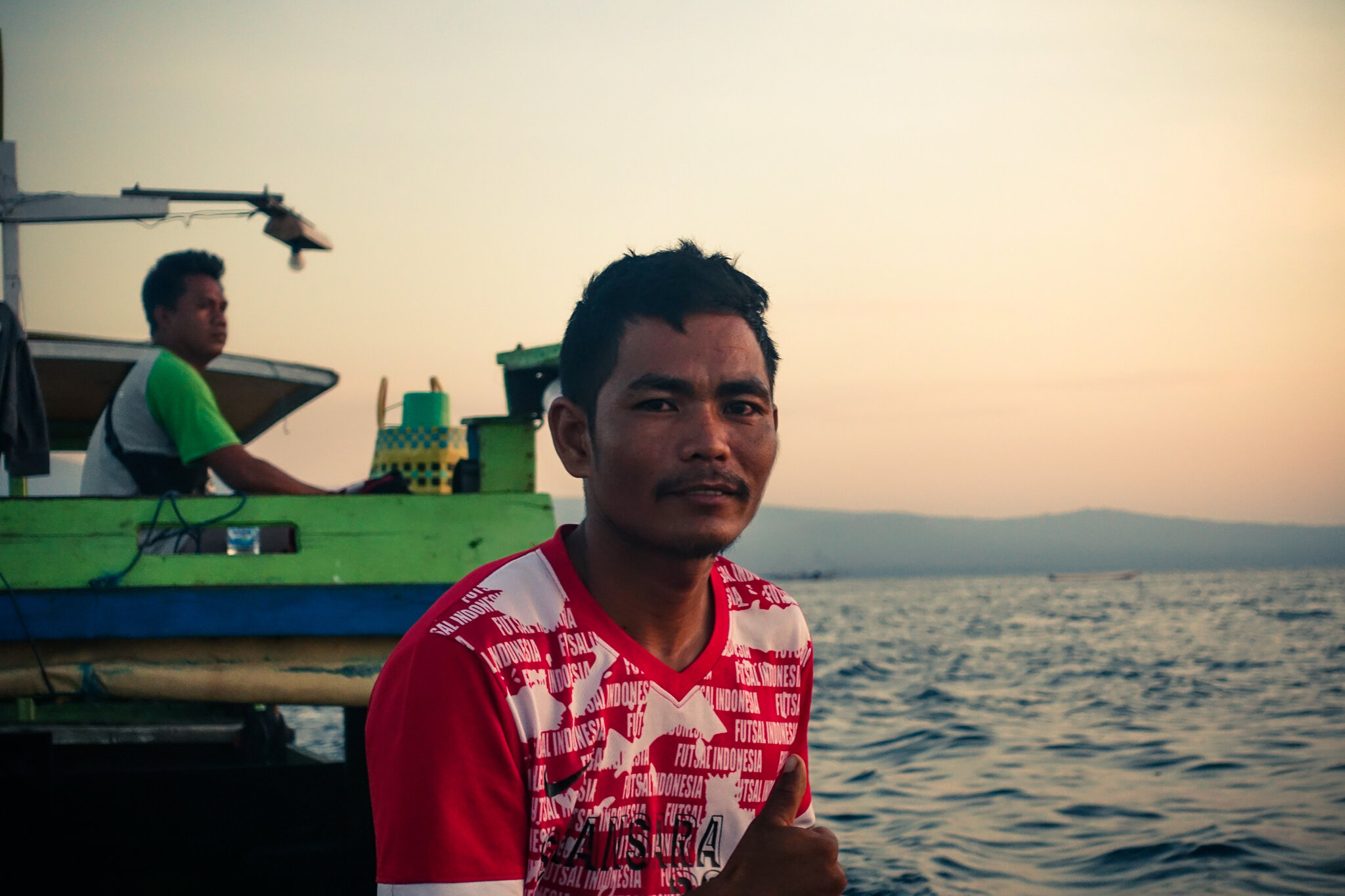 Portrait of a young Indonesian boat crew member in a red shirt at dawn on Saleh Bay, a second crew member at the tiller behind him