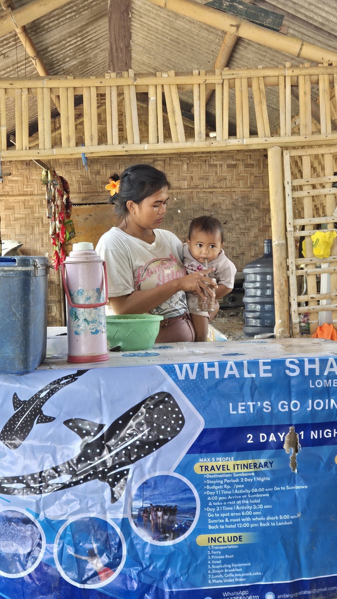 Une mère et son enfant à un stand de cuisine de rue à Labuan Jambu, Sumbawa, avec une banderole de sortie requin-baleine derrière eux
