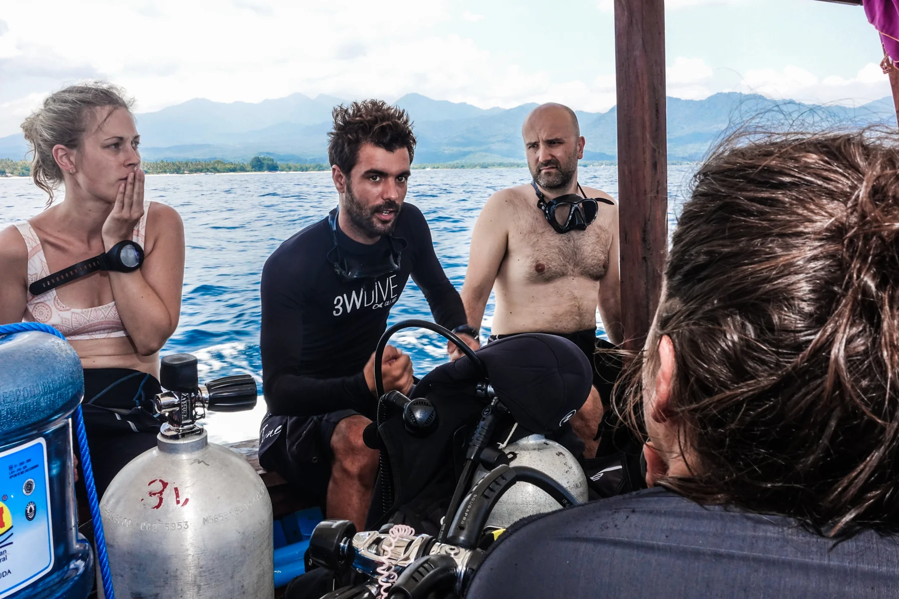 Dive guide briefing three divers on a wooden dive boat with Lombok's mountains behind, one diver checking her dive computer