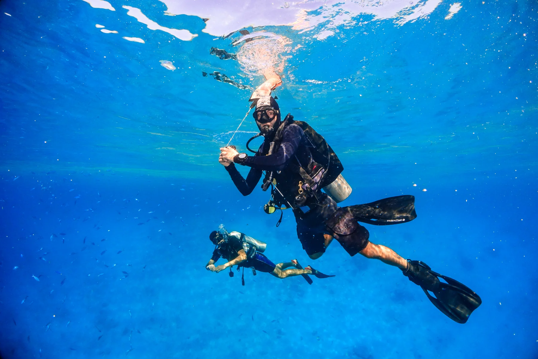 Scuba diver deploying a surface marker buoy on a reel line toward the surface in blue water, another diver below