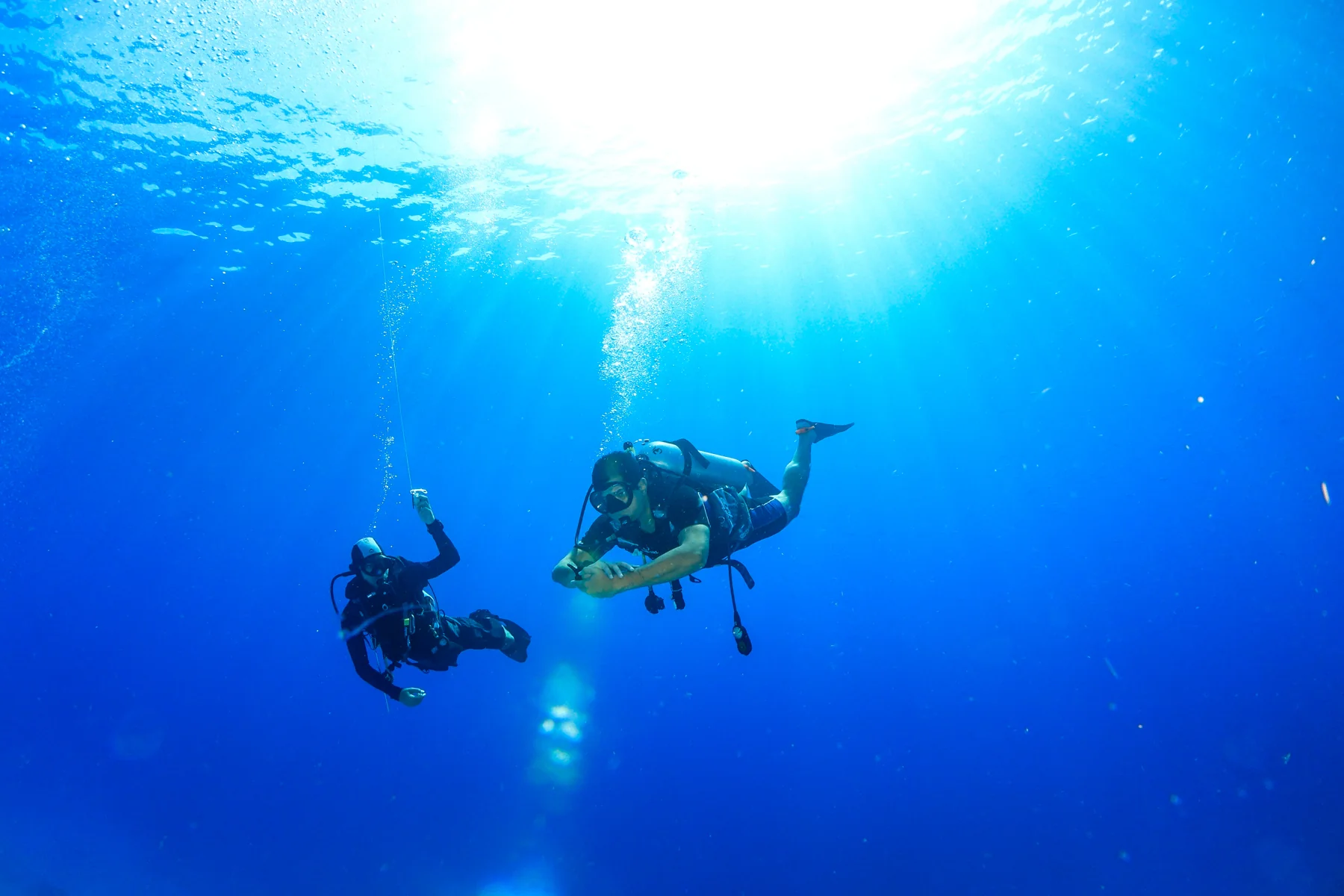 Two scuba divers hanging in deep blue water beneath the sun, one shining a torch, off Bali