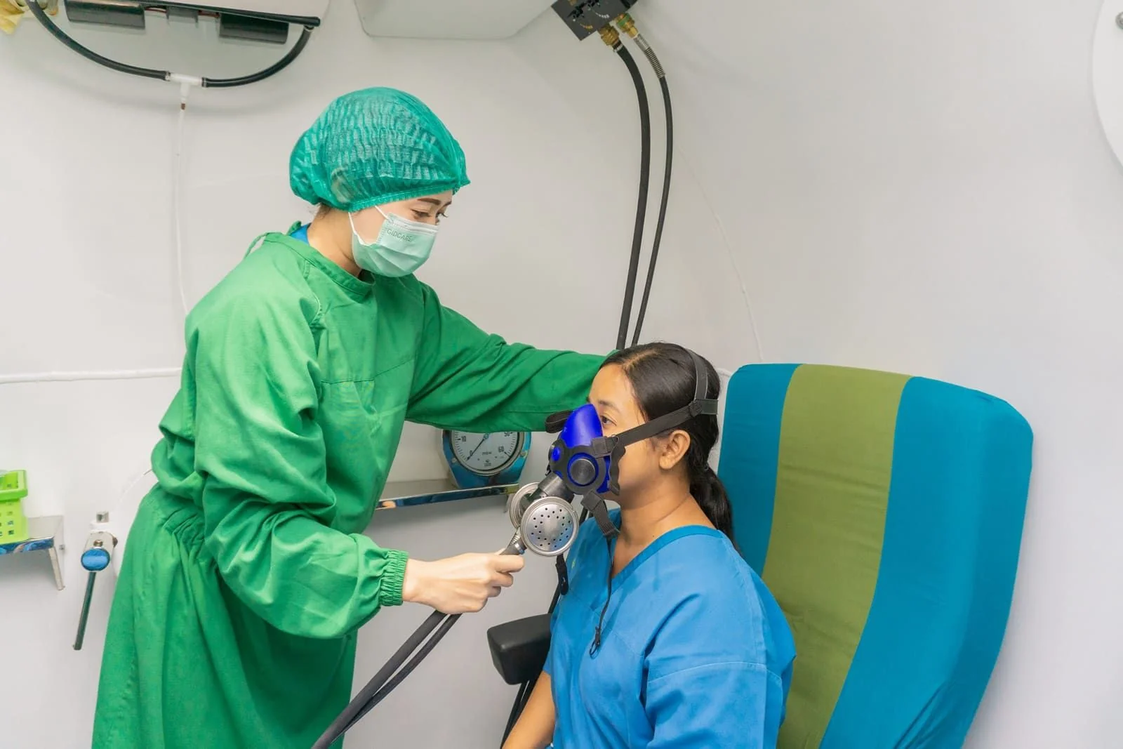 Nurse in green scrubs fitting an oxygen mask on a patient seated inside the hyperbaric chamber at Kasih Ibu Hospital Saba, Bali