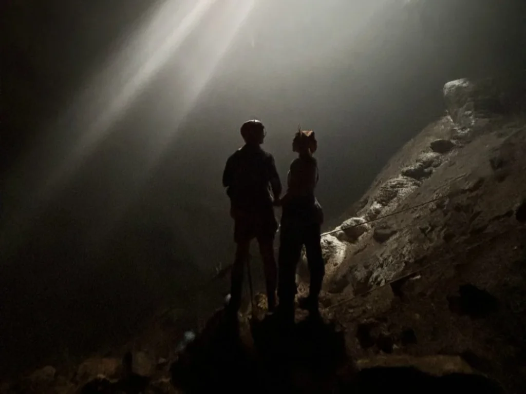 Two visitors standing in the shaft of light inside Jomblang Cave
