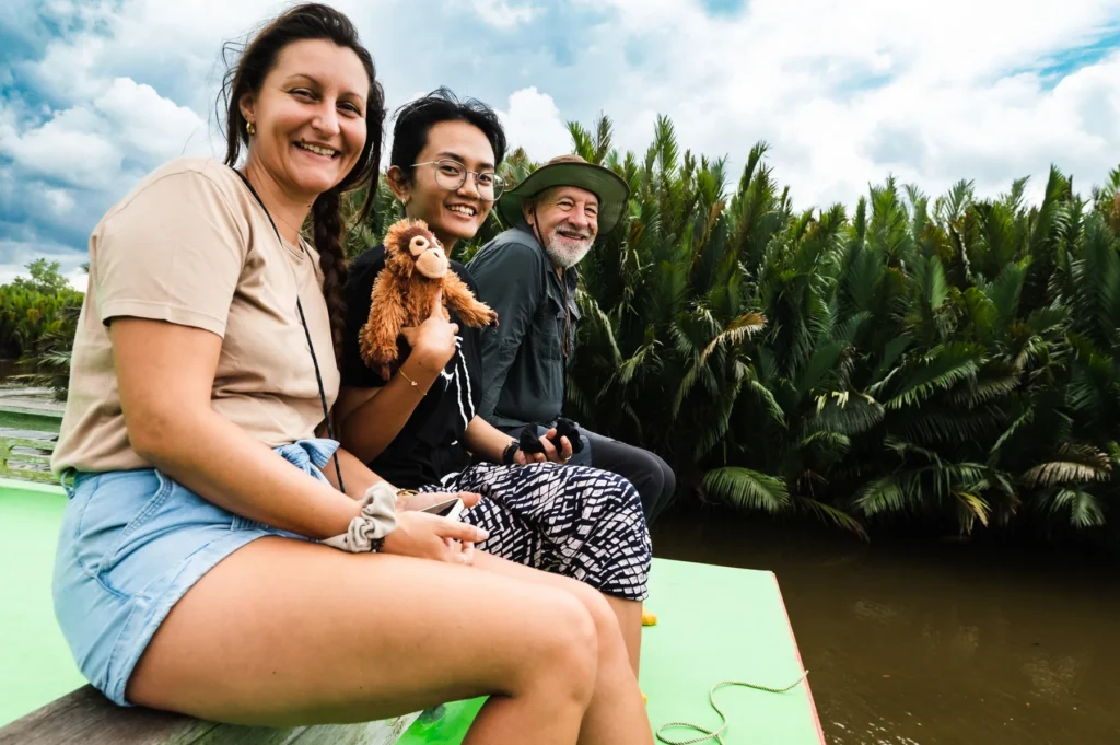 Three guests smiling on the deck of a klotok, nipa palms along the Sekonyer River behind them