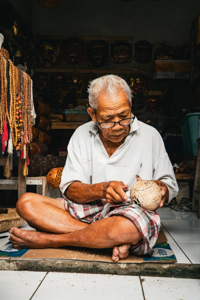 Elderly Balinese craftsman sitting cross-legged in his workshop, carving a coconut shell