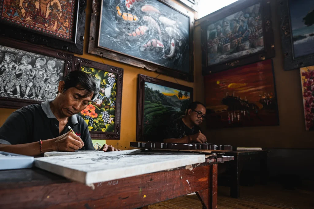 Balinese painter working on a detailed ink painting in a gallery hung with his paintings