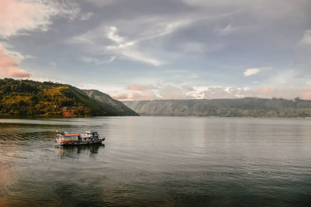 A wooden passenger boat crossing calm Lake Toba in the morning, below the caldera cliffs